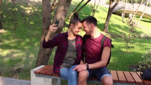 Junges verliebtes Paar wird auf einer Bank im Park sitzend fotografiert. Sie lächeln und umarmen sich. — Stockvideo