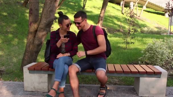 Pareja joven enamorada es fotografiada sentada en un banco en el parque.Están sonriendo y abrazándose . — Vídeos de Stock