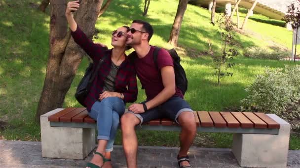 Pareja joven enamorada es fotografiada sentada en un banco en el parque.Están sonriendo y abrazándose . — Vídeo de stock
