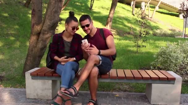 Pareja enamorada mostrar fotos en sus teléfonos inteligentes, sentado en un banco del parque . — Vídeos de Stock