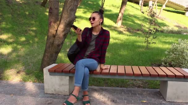 Joven chica hermosa escuchando música en su teléfono inteligente, canta y baila mientras se sienta en un banco del parque . — Vídeos de Stock
