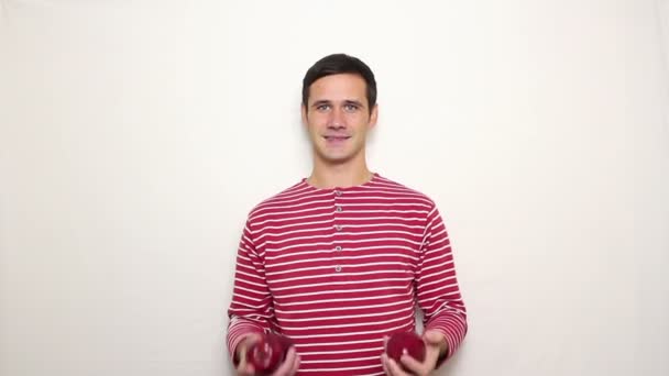 Młody przystojny facet w paski swetrze uśmiecha się szeroko i żongluje trzy czerwone jabłka. — Wideo stockowe