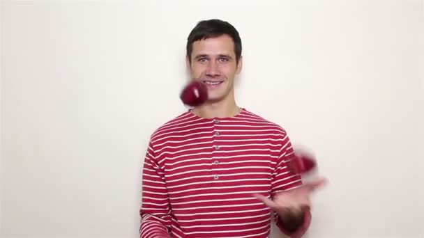 Young handsome guy in a striped sweater smiles broadly and juggles with three red apples. — Stock Video