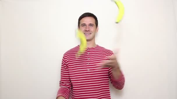 Young handsome guy in a striped sweater smiles broadly and juggles with three ripe bananas. — Stock Video