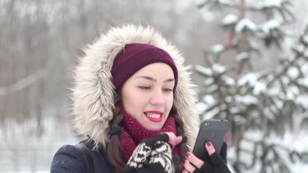 Schöne junge Mädchen blättert mit ihrem Smartphone durch soziale Netzwerke, während sie in einem Winterpark spaziert. — Stockvideo
