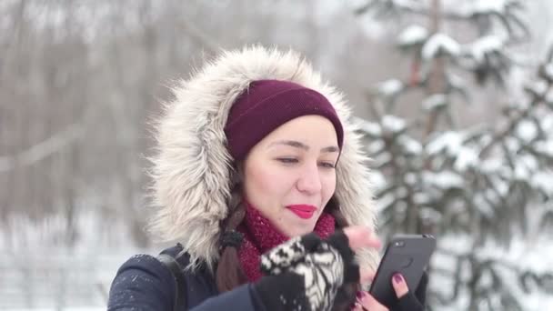 Beautiful young girl is actively flipping through the news feed on her smartphone while walking in a winter park. — Stock Video