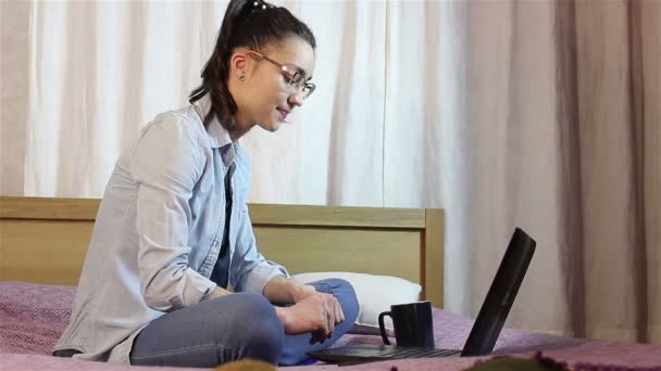 Молода приваблива дівчина працює з ноутбука, сидячи на ліжку вдома . — стокове відео