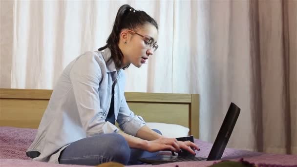 Молода приваблива дівчина працює з ноутбука, сидячи на ліжку вдома . — стокове відео