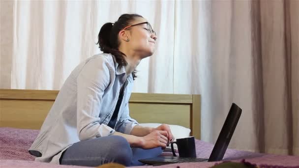 Uma jovem garota atraente está enviando uma mensagem de seu laptop, sentada na cama em casa . — Vídeo de Stock