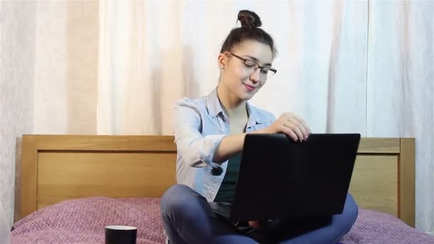Een mooi jong meisje opent haar laptop en typeing op het toetsenbord, zittend op het bed. — Stockvideo