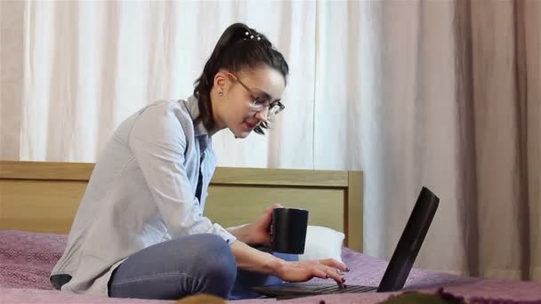 Ein junges attraktives Mädchen schreibt eine SMS von ihrem Laptop, zu Hause auf dem Bett sitzend. — Stockvideo