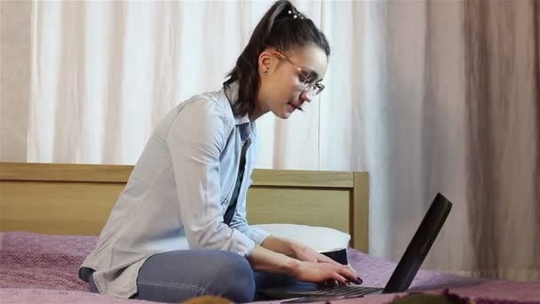 Молода приваблива дівчина пише повідомлення зі свого ноутбука, сидячи на ліжку вдома . — стокове відео