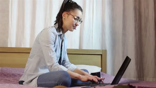 Una giovane ragazza attraente sta messaggiando un messaggio dal suo computer portatile, seduto sul letto a casa . — Video Stock