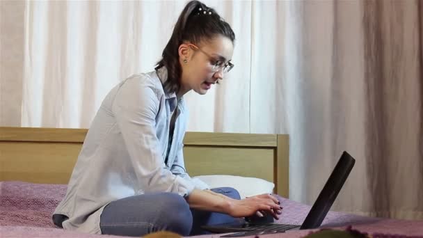 Молода приваблива дівчина працює над своїм ноутбуком, сидячи на ліжку вдома . — стокове відео
