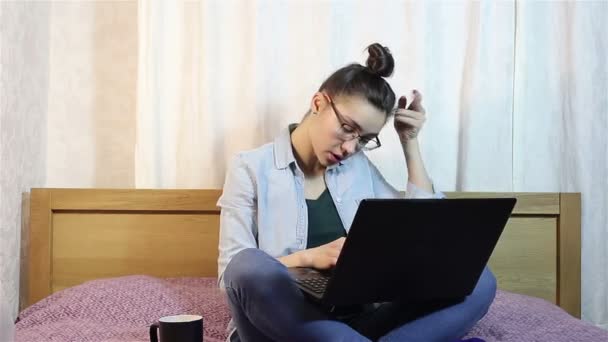 Una chica atractiva joven está enviando un mensaje de texto desde su computadora portátil, sentado en la cama en casa . — Vídeo de stock