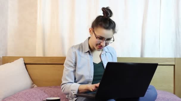 Una chica atractiva joven está enviando un mensaje de texto desde su computadora portátil, sentado en la cama en casa . — Vídeos de Stock
