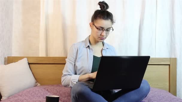 魅力的な少女は自宅のベッドの上に座って、彼女のノート パソコンから携帯メールを打って. — ストック動画