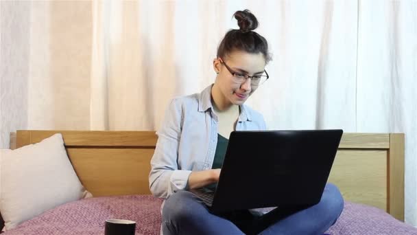 Молода приваблива дівчина пише повідомлення зі свого ноутбука, сидячи на ліжку вдома . — стокове відео