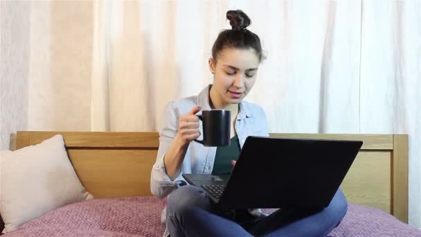 A young beautiful girl working from her laptop while drinking coffee from a black cup.