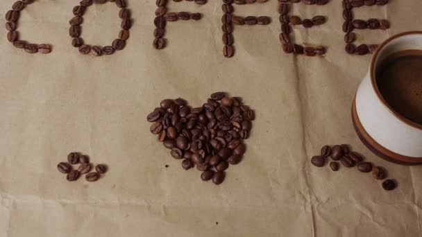 Filiżankę aromatycznej kawy umieszcza się na papierze rzemieślniczym ze słowem "kawa" wyukładane z ziaren kawy. — Wideo stockowe