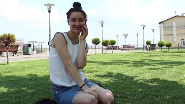 Mladá atraktivní dívka v bílém tričku mluví na telefonu, sedícího na trávě v parku. — Stock video