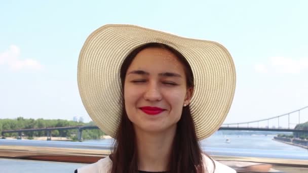 Närbild porträtt en vacker ung flicka i en vit hatt tittar in i kameran och ler. En ung attraktiv flicka i en vit hatt, höjer blicken mot himlen och vindögdhetar från solens strålar. — Stockvideo
