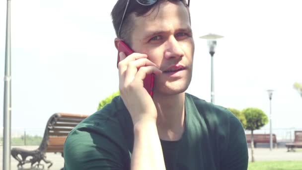 Primer plano retrato Joven en camiseta verde hablando por teléfono mientras está sentado en la hierba en el parque . — Vídeos de Stock