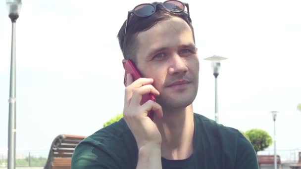 Крупным планом портрет Молодой парень в зеленой футболке разговаривает по телефону и широко улыбается, сидя на траве в парке . — стоковое видео