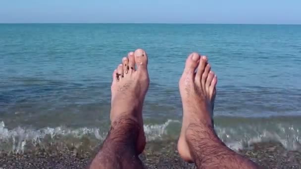 Vista de las piernas de un hombre acostado en la orilla del mar. Piernas masculinas sobre un fondo de mar azul.Un hombre disfruta de unas vacaciones en una playa de guijarros . — Vídeo de stock