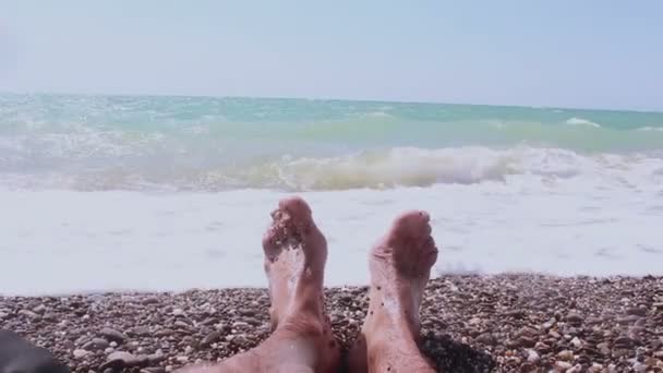 Vista de las piernas de un hombre acostado en la orilla del mar. Piernas masculinas sobre un fondo de mar azul.Un hombre disfruta de unas vacaciones en una playa de guijarros . — Vídeo de stock