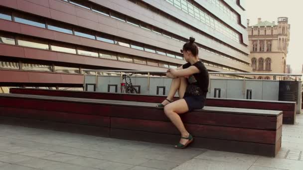 Menina bonita jovem está descansando em um banco contra o fundo de um prédio de escritórios . — Vídeo de Stock