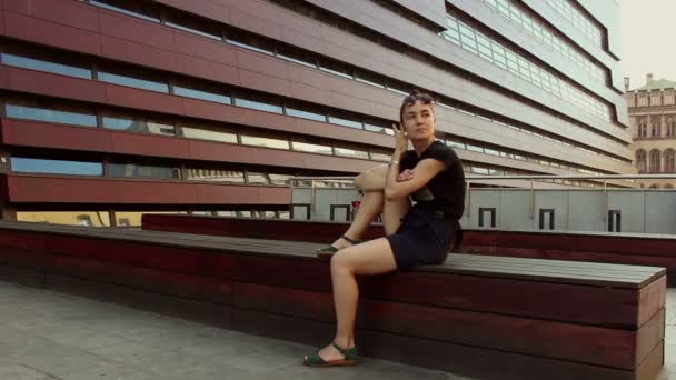 Menina bonita jovem está descansando em um banco contra o fundo de um prédio de escritórios . — Vídeo de Stock