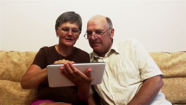 Yaşlı bir çift evdeki bir kanepede oturuyor ve tabletteki fotoğrafları izliyor, gülümsüyor ve aktif bir şekilde konuşuyor.. — Stok video