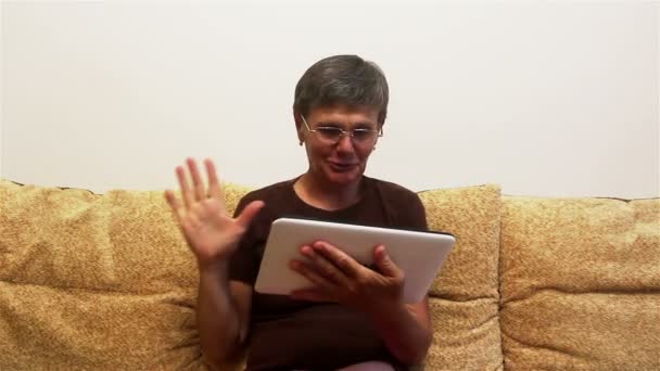 Uma mulher adulta atraente é vídeo conversando com sua família, em um tablet pc, sentado em um sofá em casa . — Vídeo de Stock