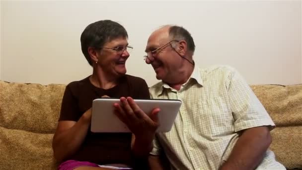 Una pareja de ancianos está sentada en un sofá en casa y viendo fotos en una tableta PC, sonriendo y hablando activamente . — Vídeos de Stock