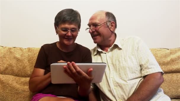 Пожилая пара сидит дома на диване и смотрит фотографии на планшете ПК, улыбаясь и активно разговаривая . — стоковое видео