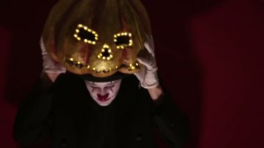 Palyaço makyajındaki korkunç adam Cadılar Bayramı için bir balkabağı tutuyor ve kafasına takıyor. Korkunç palyaço kafasına balkabağı şeklinde bir maske takıyor. Korkunç palyaço elinde parlak gözlü ve ağzı olan bir balkabağı tutuyor..