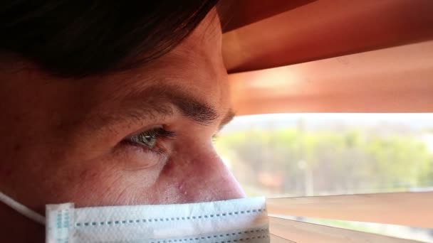 Mladý pohledný muž v lékařské masce se dívá okenními žaluziemi. Mladý muž v lékařské masce se dívá oknem, je na izolaci.. — Stock video