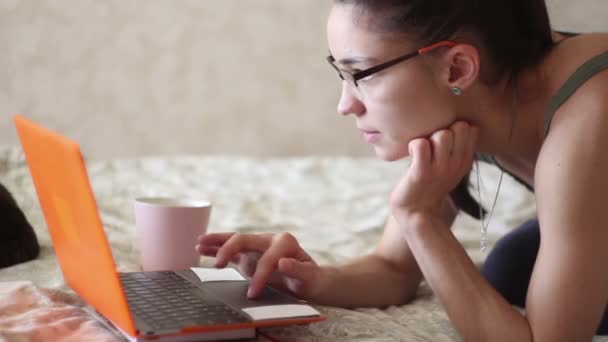 Joven chica atractiva está trabajando en su computadora portátil, acostado en la cama en casa. Una joven linda chica está acostada en la cama y escribiendo en su computadora portátil . — Vídeo de stock