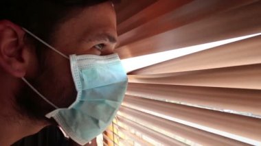 Tıbbi maske takmış genç yakışıklı bir adam pencere perdelerinden içeri bakıyor. Tıbbi maskeli genç bir adam pencereden bakar, kendini izole eder..