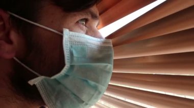 Tıbbi maske takmış genç yakışıklı bir adam pencere perdelerinden içeri bakıyor. Tıbbi maskeli genç bir adam pencereden bakar, kendini izole eder..