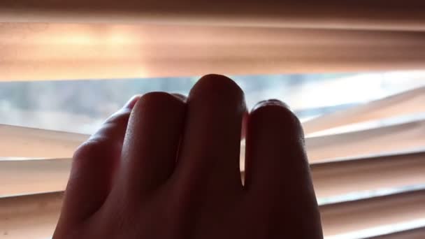 En ung stilig man öppnar persiennerna med handen och tittar ut genom fönstret.. — Stockvideo