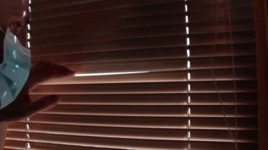 Tıbbi maske takmış genç yakışıklı bir adam pencere perdelerinden içeri bakıyor. Tıbbi maskeli genç bir adam pencereden bakar, kendini izole eder..