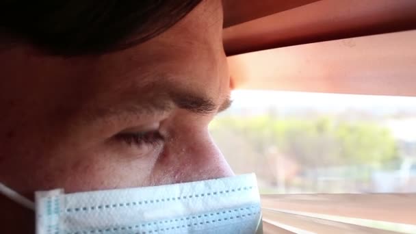 Tıbbi maske takmış genç yakışıklı bir adam pencere perdelerinden içeri bakıyor. Tıbbi maskeli genç bir adam pencereden bakar, kendini izole eder.. — Stok video