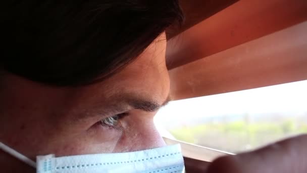 Een jonge knappe man met een medisch gezichtsmasker kijkt door de gordijnen. Een jongeman met een medisch masker kijkt door het raam, op zelfisolatie.. — Stockvideo