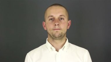 Beyaz tişörtlü genç yakışıklı adam, hoşça kal deyip elini sallıyor. Gri arkaplanda bir portre.