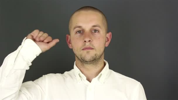 Jonge knappe man in een wit shirt, neemt afscheid en zwaait met zijn hand. Portret op een grijze achtergrond. — Stockvideo