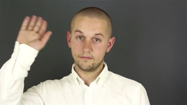Jonge knappe man in een wit shirt, neemt afscheid en zwaait met zijn hand. Portret op een grijze achtergrond. — Stockvideo