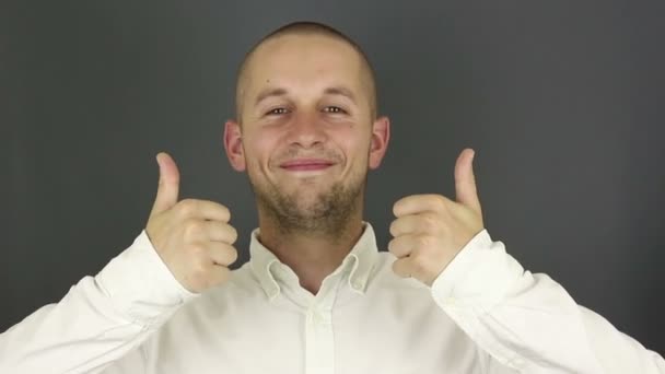 Divertido chico guapo sonríe y muestra el pulgar hacia arriba con ambas manos. Retrato sobre fondo gris . — Vídeos de Stock