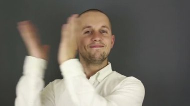 Komik yakışıklı bir adam coşkusunu aktif olarak ifade ediyor ve ellerini çırpıyor. Genç bir adamın portresi, coşkusunu dile getiriyor ve alkışlıyor. Gri arkaplanda bir portre.